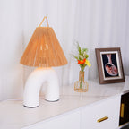 Low angle on the white arched ceramic base and woven rope shade of this Clérachic lamp, beside a posy and a framed photo.