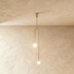 Clérachic slim arch pendant in gold with two glowing frosted globes at different heights, against limewashed plaster.