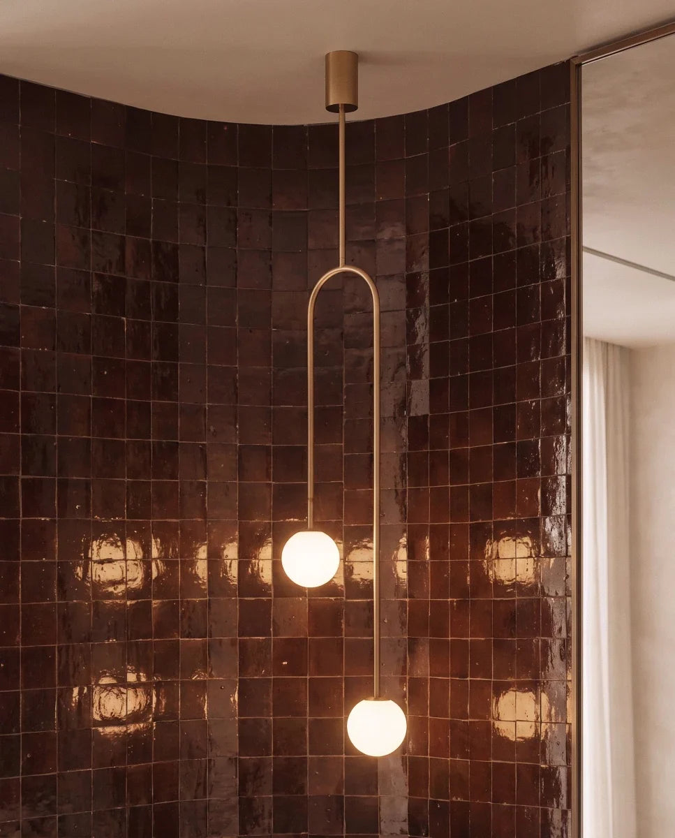 Clérachic gold arch pendant with two glowing globes against a curved wall of glossy brown tiles, seen close up.