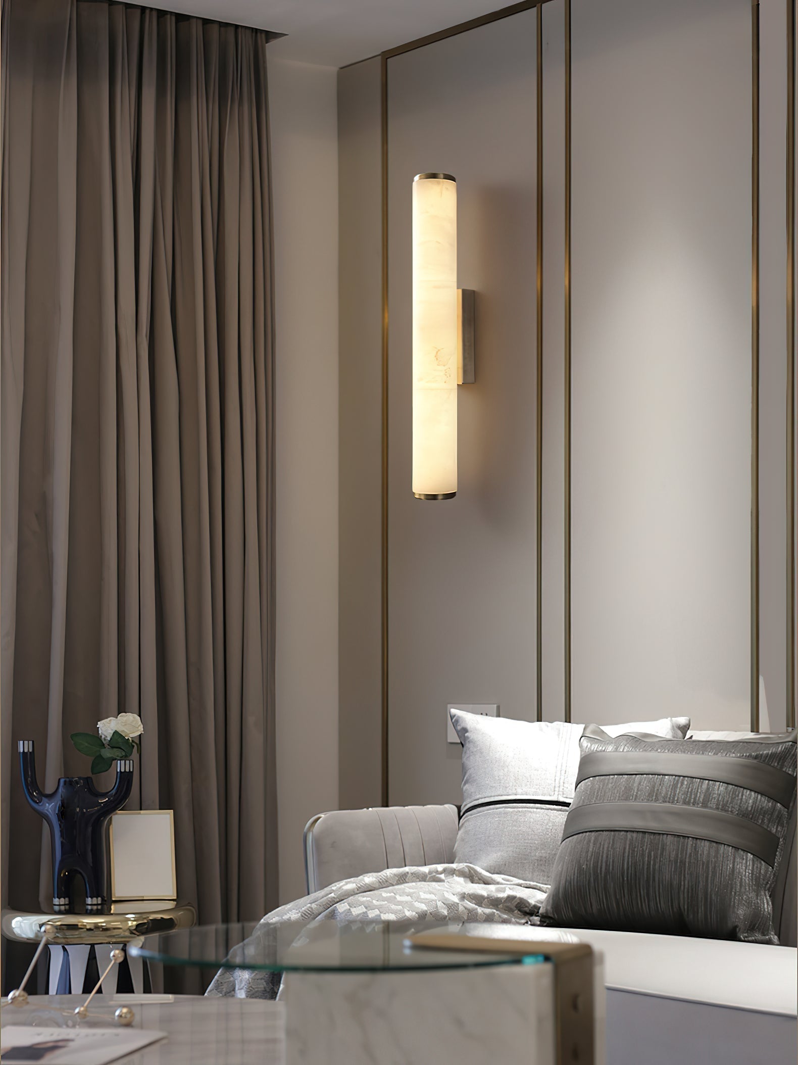 The Clérachic sconce in brass beside a bed, throwing warm light across ribbed wall panels and floor-length curtains.