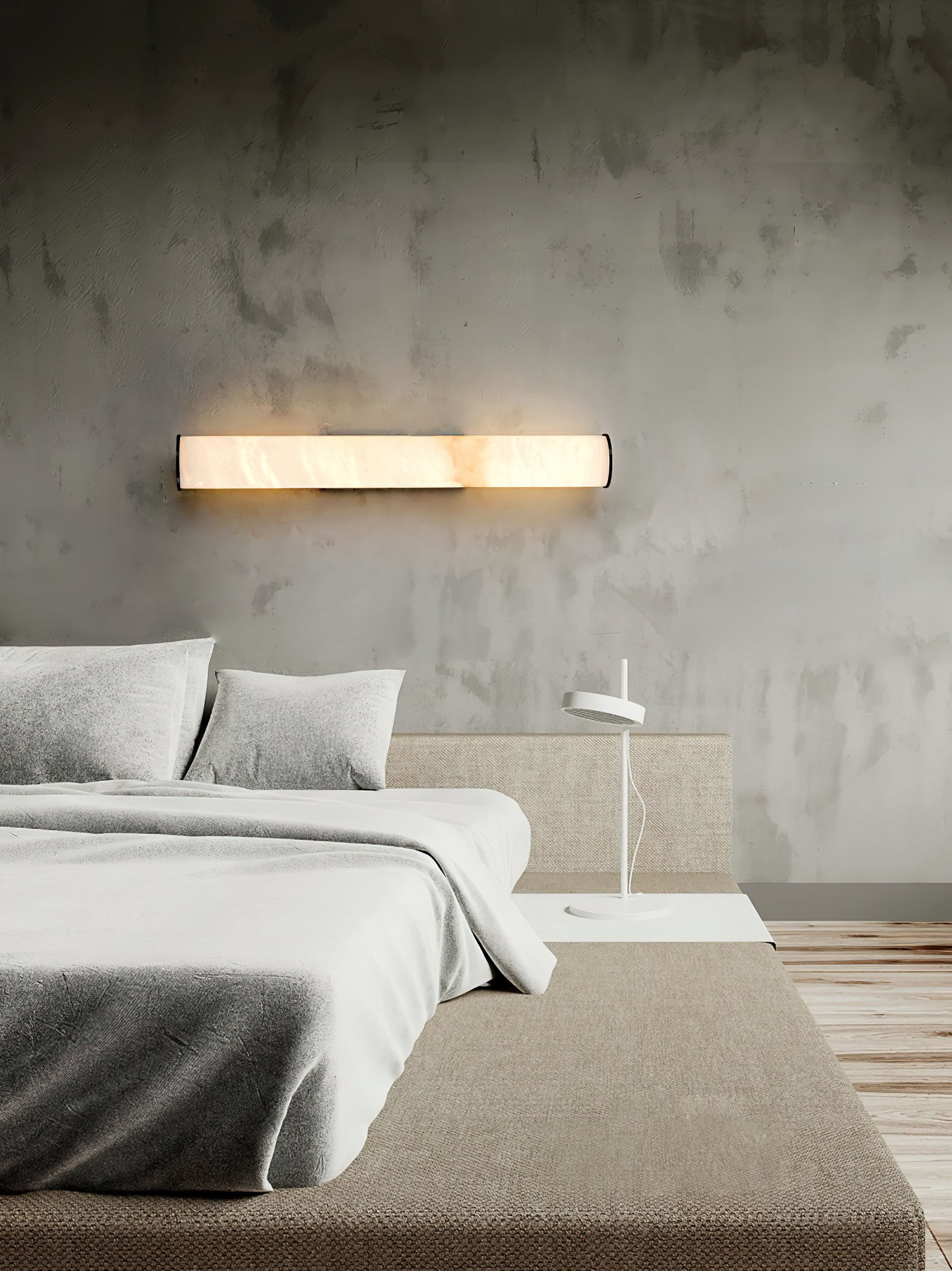 The Clérachic sconce fitted horizontally on a raw concrete wall, glowing above a bed made up in grey linen.