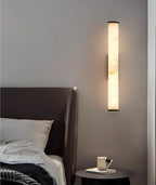 The Clérachic sconce above a round nightstand, alabaster veining lit beside a leather-wrapped headboard.