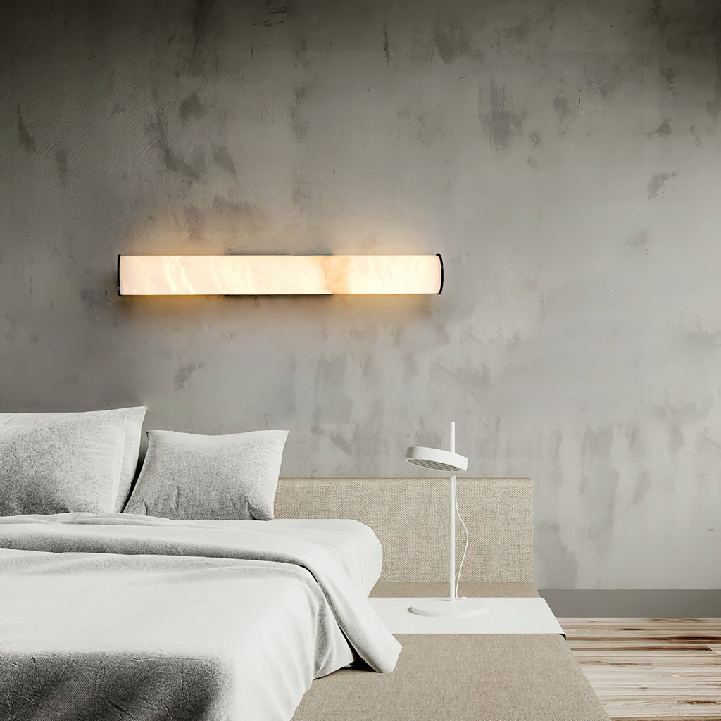 Clérachic sconce fitted horizontally over a headboard, its alabaster bar glowing against textured concrete.