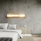 Clérachic sconce fitted horizontally over a headboard, its alabaster bar glowing against textured concrete.
