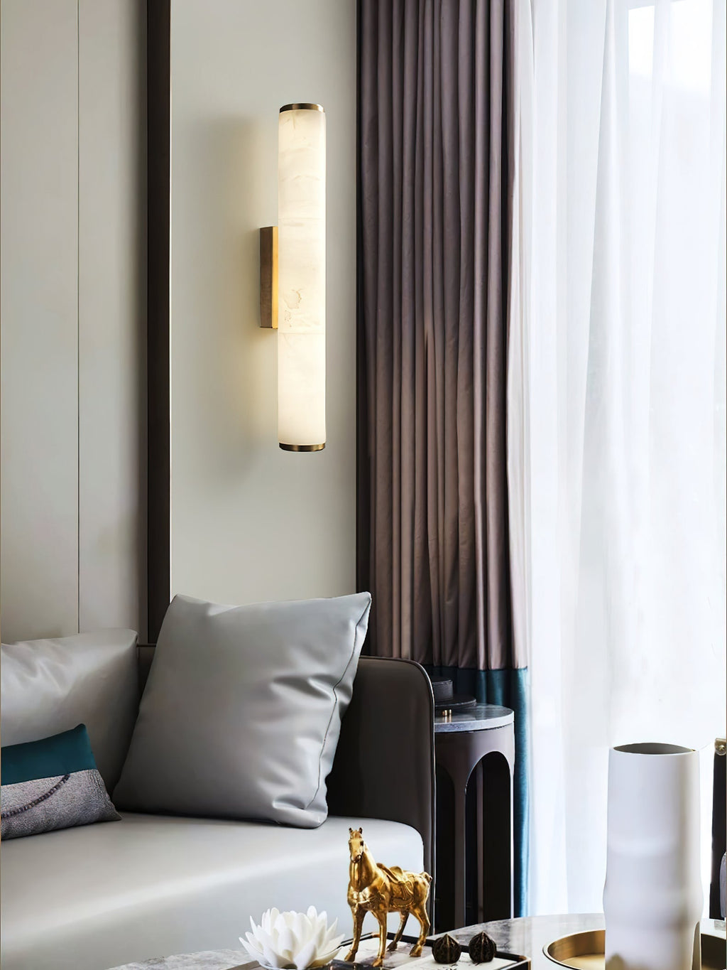 The Clérachic sconce beside heavy brown curtains, lighting a pale sofa, brass ornament and ceramic vase.