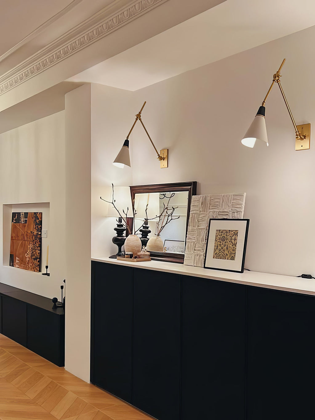 Two Clérachic articulated wall lights on a cream wall above a black sideboard, with ceramics and framed prints below and a herringbone floor.