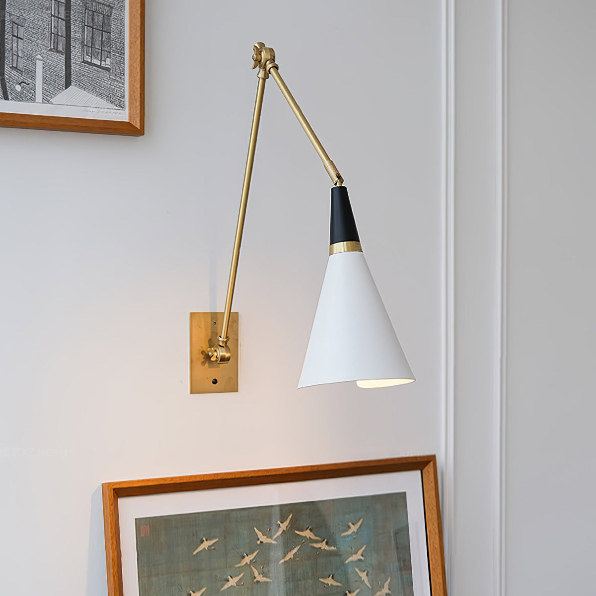 Clérachic wall light with a white cone shade on a folded brass arm, above a framed painting of birds on a pale wall.