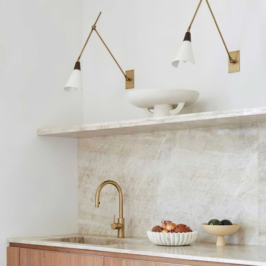 Two Clérachic wall lights with white cone shades and brass arms above a marble kitchen shelf, tap and wooden counter.