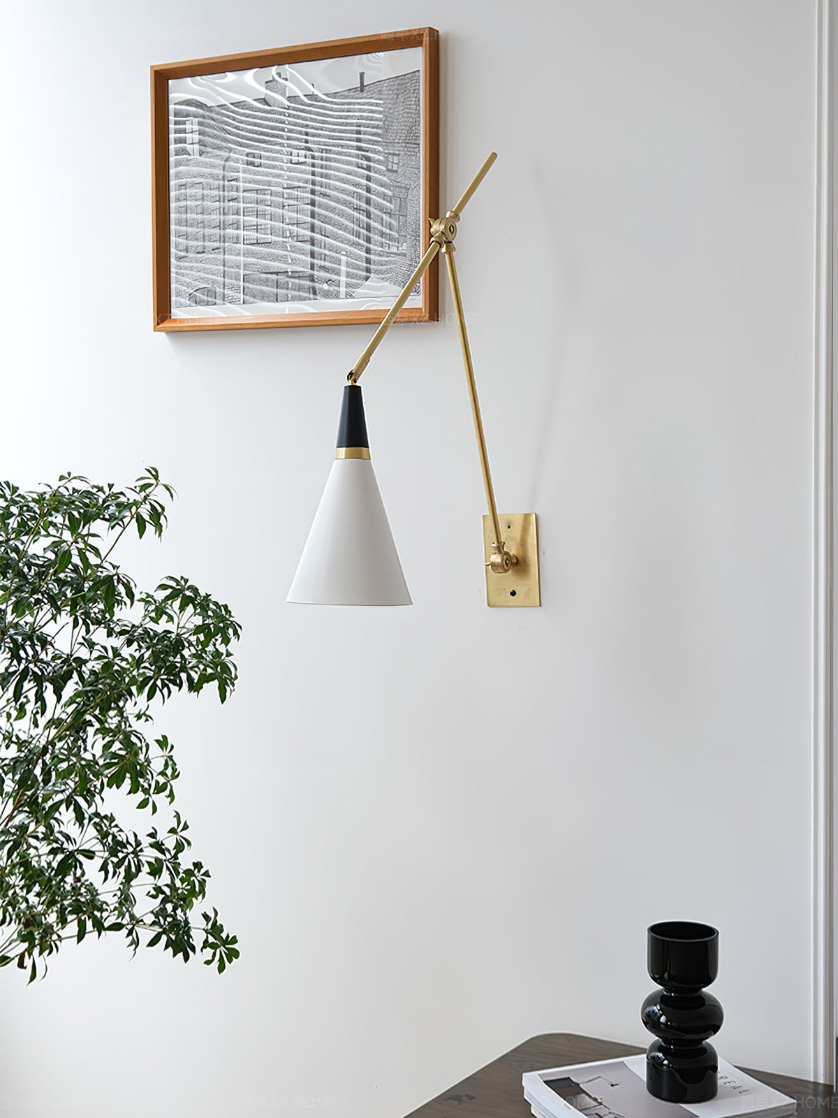 Clérachic wall light with a white cone shade on a brass arm, beside a framed print and a leafy branch, on a white wall.