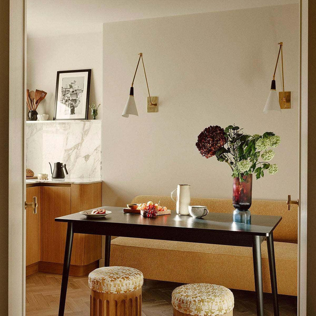 Two Clérachic wall lights on a cream wall above a dining table with a vase of dark flowers, in a corner with marble shelving.