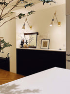 Two Clérachic wall lights with white cone shades above a black cabinet, framed prints and a branch in the foreground of a cream kitchen.