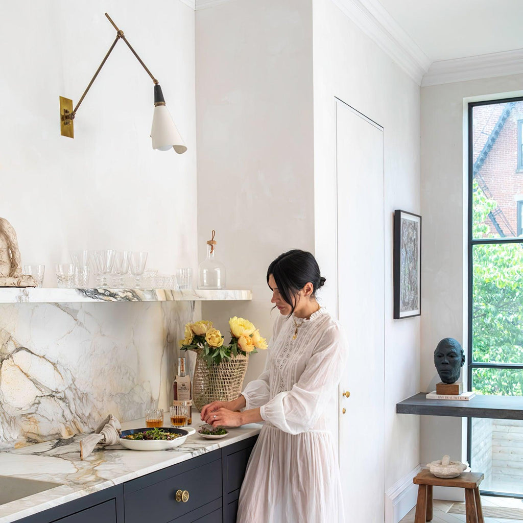 Clérachic wall light on a limewashed wall above a marble kitchen counter, with a woman in a white dress preparing flowers and food.