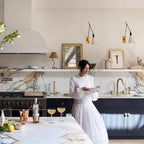Two Clérachic wall lights above a marble kitchen shelf, with a gilt mirror, framed prints and a woman carrying a plate below.