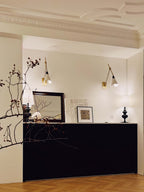 Two Clérachic wall lights on a cream wall with ornate ceiling mouldings, above a black cabinet and a bare branch in a vase.