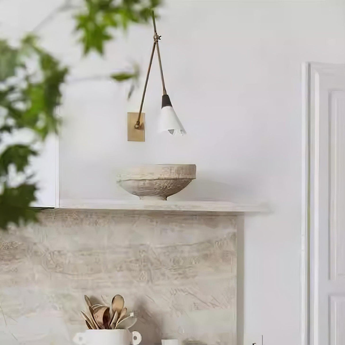 Clérachic wall light with a white cone shade at the end of a brass arm, set above a stone mantel and bowl in a pale room.