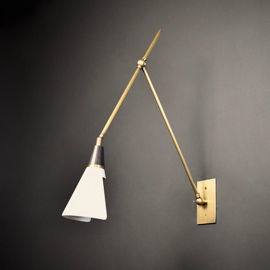 Clérachic wall light with a white cone shade and brass arms on a dark grey wall, the arms making a wide V from the backplate.
