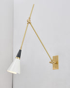 Clérachic wall light with a white cone shade tilted down, hung from a brass arm on a pale grey wall beside a corner.