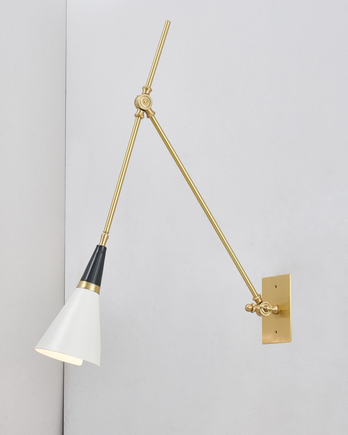 Clérachic wall light with a white cone shade tilted down, hung from a brass arm on a pale grey wall beside a corner.