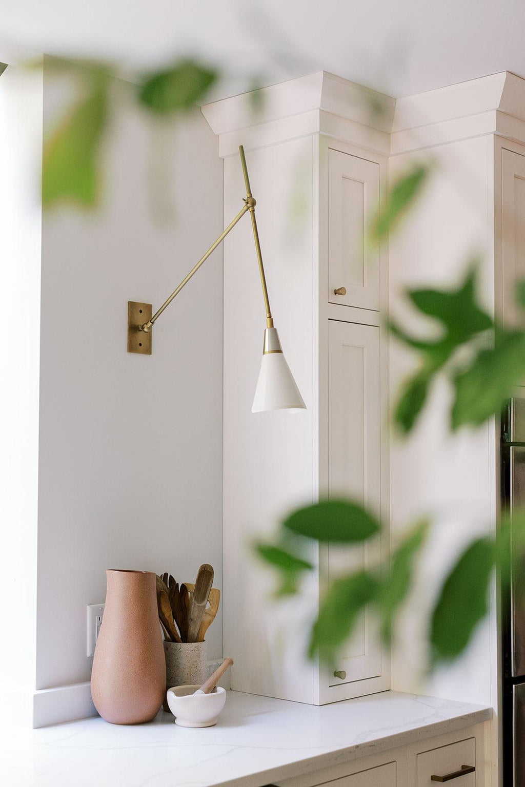 Clérachic wall light with a white cone shade on a brass arm, beside a blurred green plant and tall white kitchen cabinets.