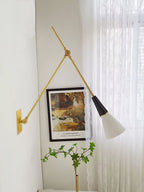Clérachic wall light with a wide arm angle beside lace curtains, its white cone shade lit from inside beside a framed garden print.