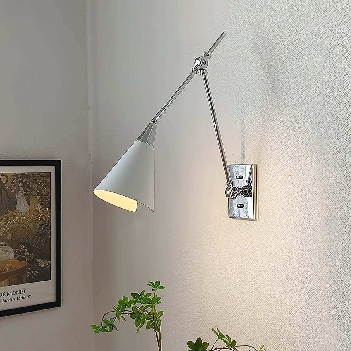 Clérachic wall light in bright nickel, with a white cone shade lit inside, beside a framed garden print on a pale wall.