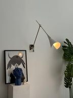 Clérachic wall light in nickel with its white cone shade lit, above a framed black-and-white portrait and a blue sculpture on a white plinth.