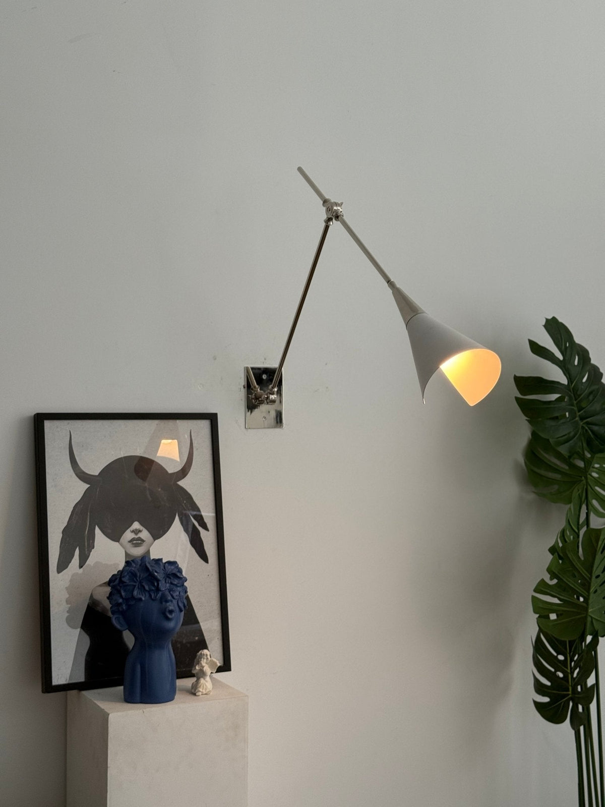 Clérachic wall light in nickel with its white cone shade lit, above a framed black-and-white portrait and a blue sculpture on a white plinth.