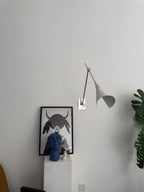 Clérachic wall light in nickel with a white cone shade, hung on a pale wall above a framed portrait and a plinth, a green leaf at the right.