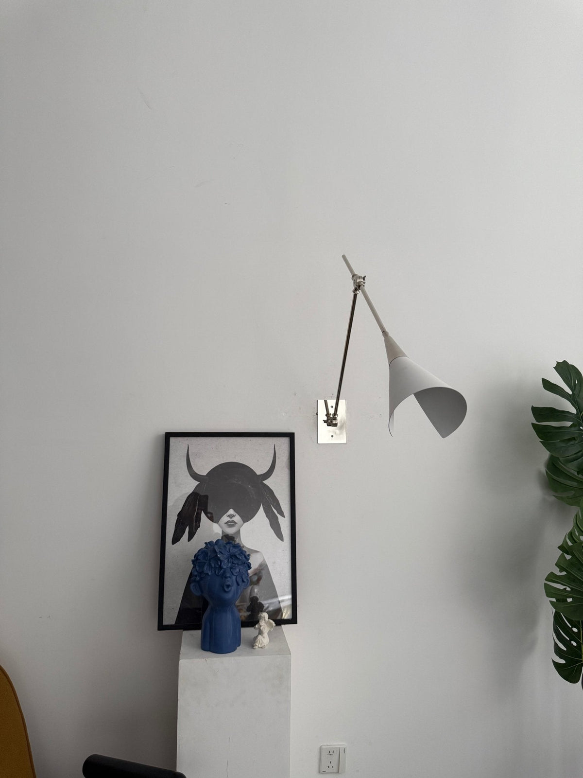 Clérachic wall light in nickel with a white cone shade, hung on a pale wall above a framed portrait and a plinth, a green leaf at the right.