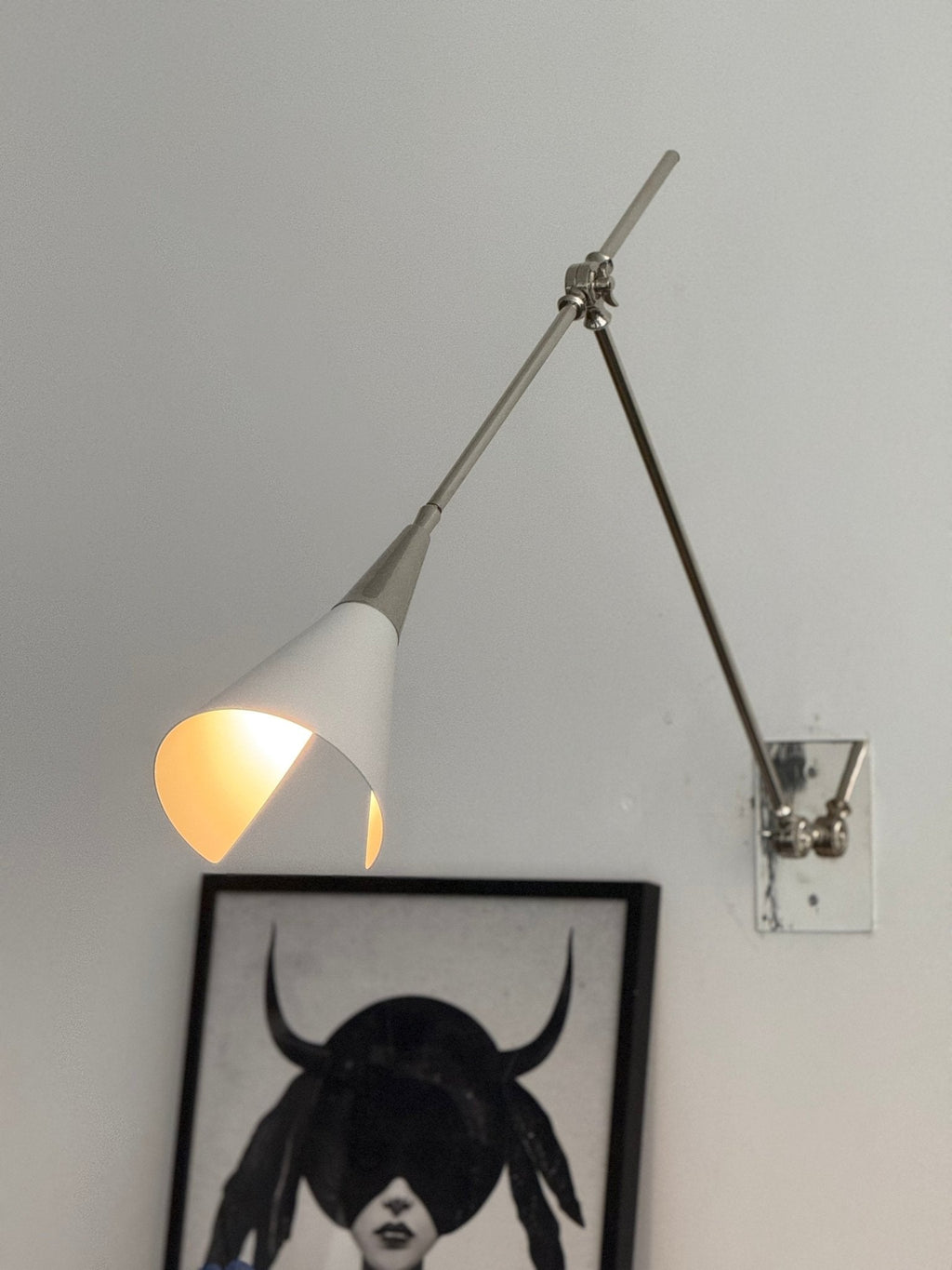 Clérachic wall light in nickel, its white cone shade lit inside, beside a framed portrait with a blue ceramic head on a plinth below.