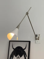 Clérachic wall light in nickel, its white cone shade lit inside, beside a framed portrait with a blue ceramic head on a plinth below.