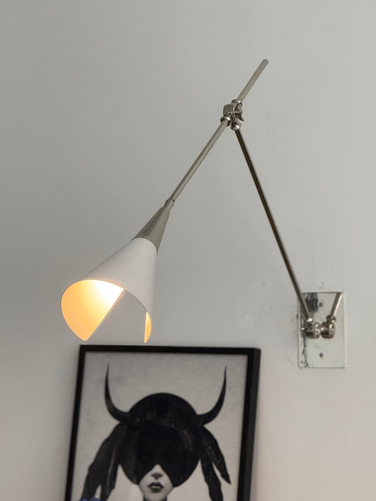 Clérachic wall light in nickel, its white cone shade lit inside, beside a framed portrait with a blue ceramic head on a plinth below.