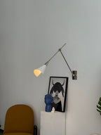 Clérachic wall light in nickel with a lit white cone shade, above a framed portrait and a yellow chair on a pale wall.