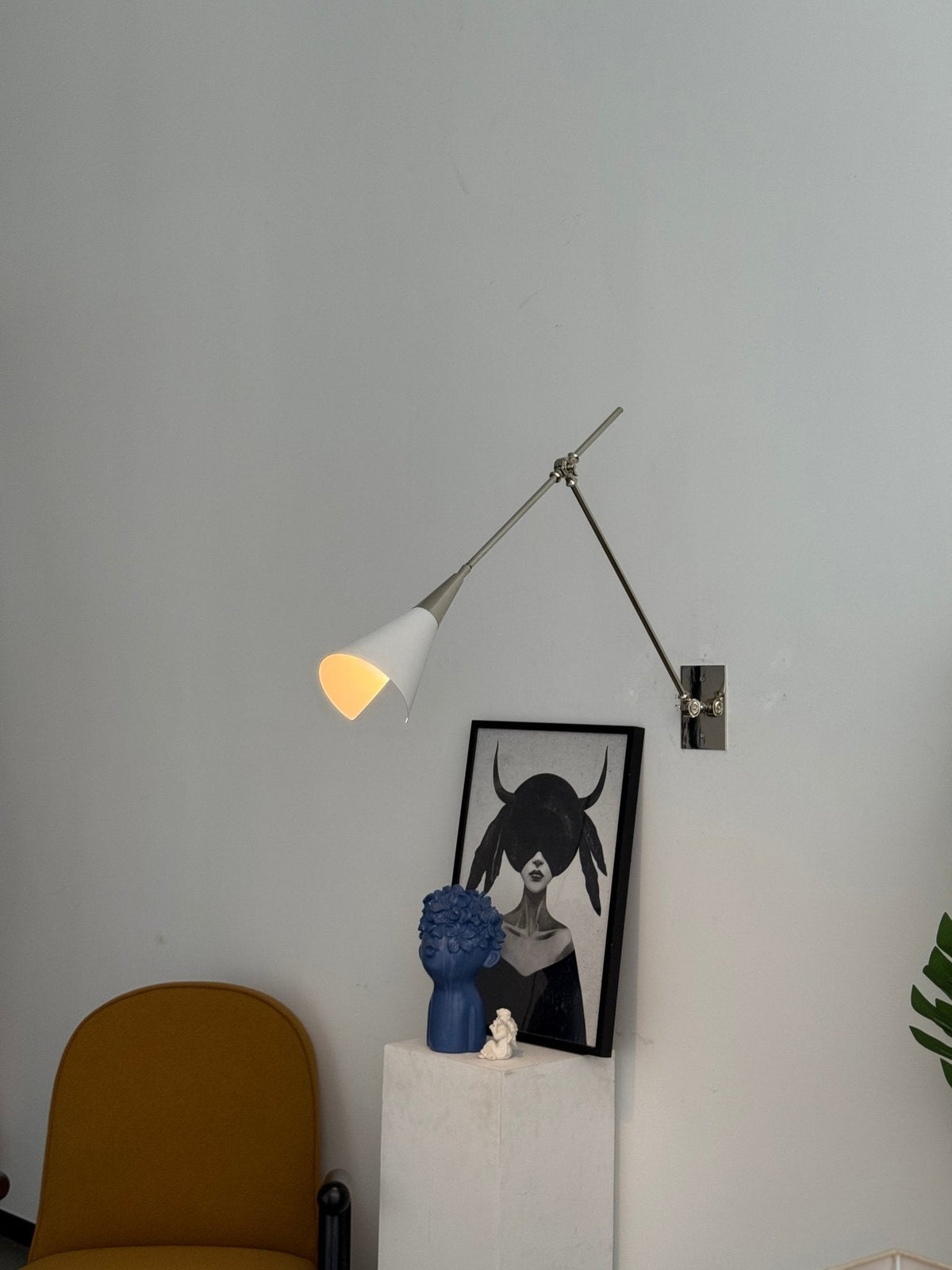 Clérachic wall light in nickel with a lit white cone shade, above a framed portrait and a yellow chair on a pale wall.
