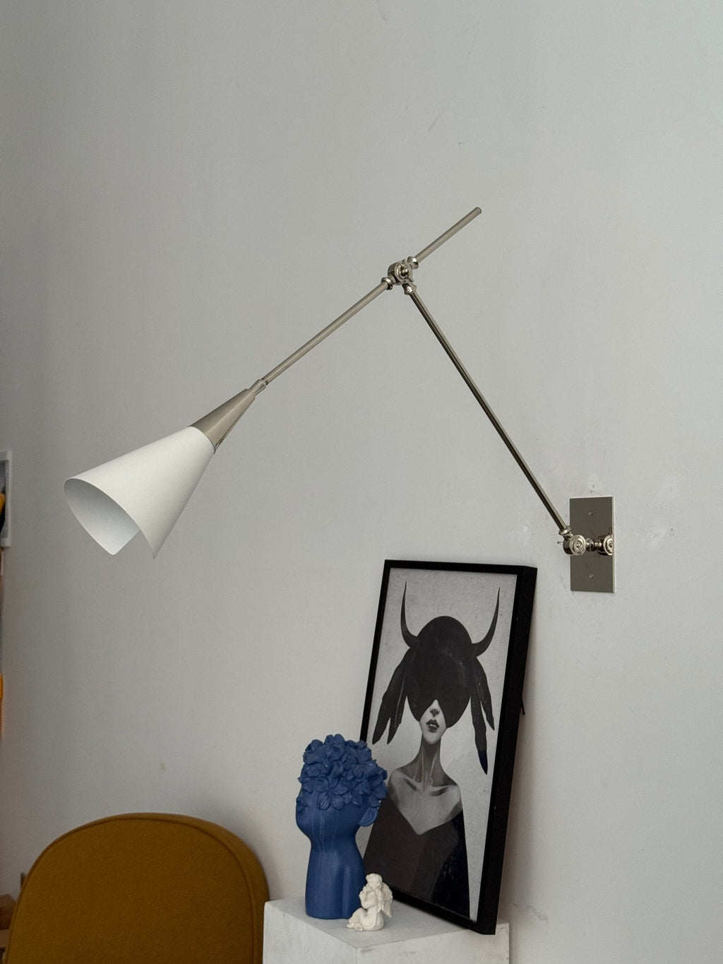 Clérachic wall light in nickel with its white cone shade unlit, on a pale wall with a framed portrait, a blue sculpture and a yellow chair below.
