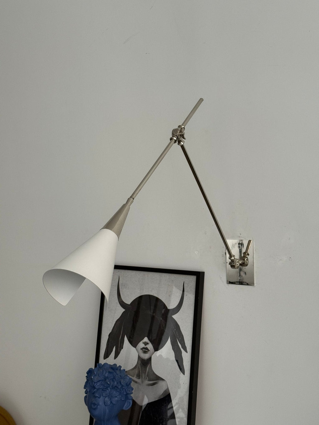 Clérachic wall light in nickel with a white cone shade tilted down, in front of a framed portrait and a blue sculpture.