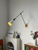 Clérachic wall light with a black neck and brass arms lit above a white arched shelf with a white framed print and dried flowers, on a pale wall.