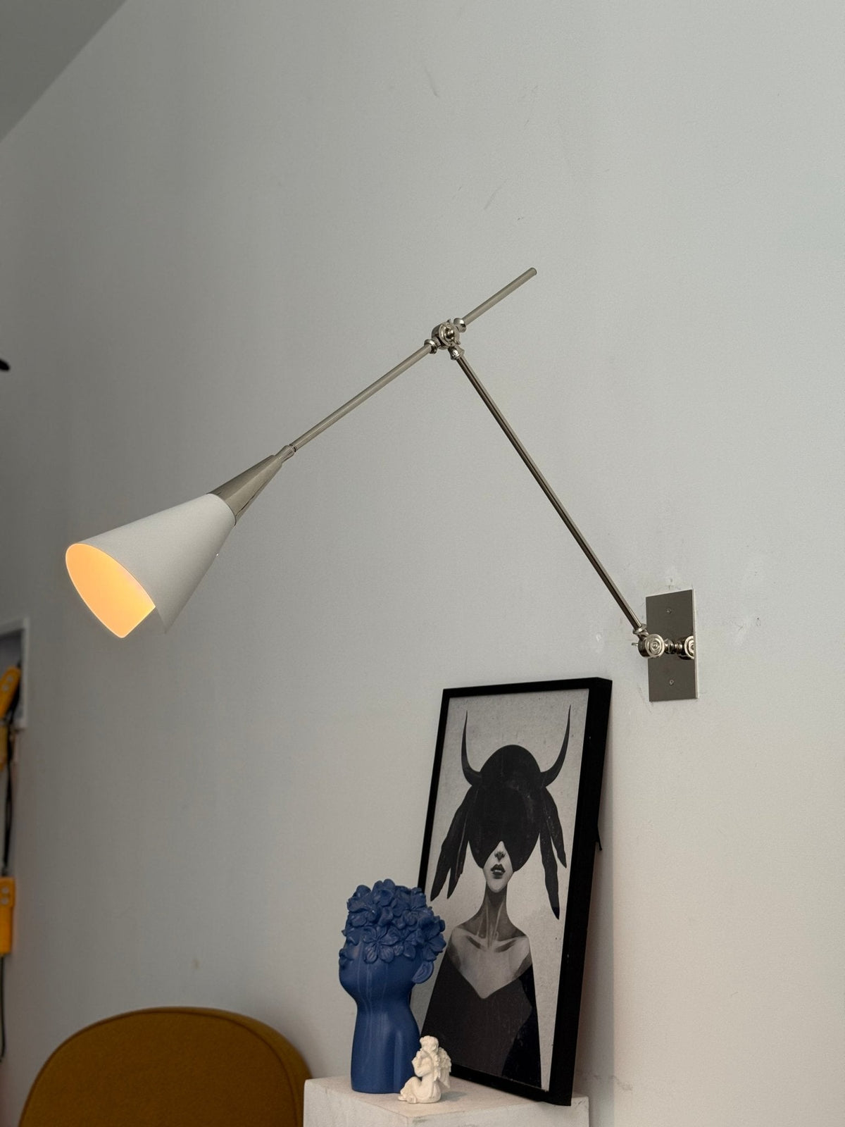 Clérachic wall light in nickel with a lit white cone shade, above a framed portrait and a blue ceramic head, with a yellow chair below.