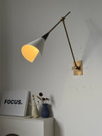 Clérachic wall light with a lit white cone shade glowing warm, on a brass arm above a white shelf with a white framed print and a blue vase.