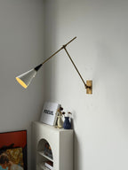 Clérachic wall light with a lit white cone shade on a brass arm and backplate, above a white arched unit with a small white print.