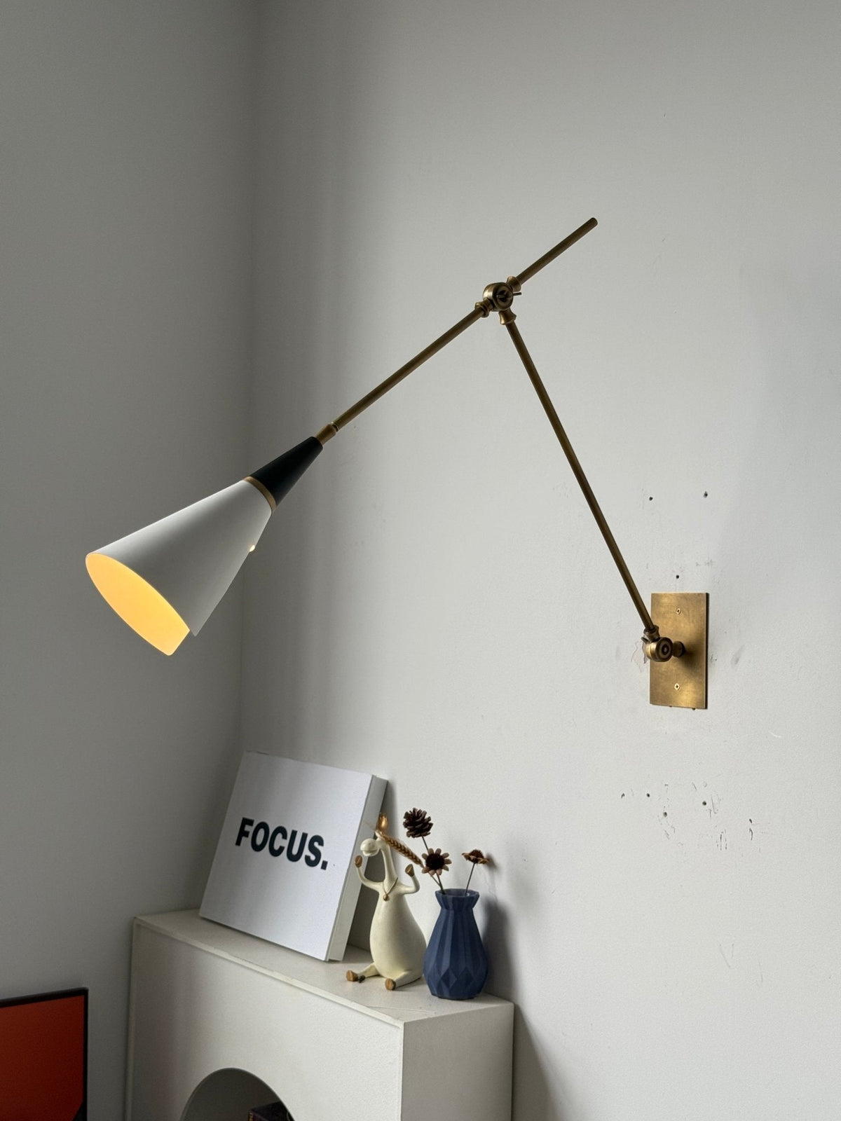 Clérachic wall light with a white cone shade lit warmly and a black neck, on long brass arms above a white arched shelf and a white print.