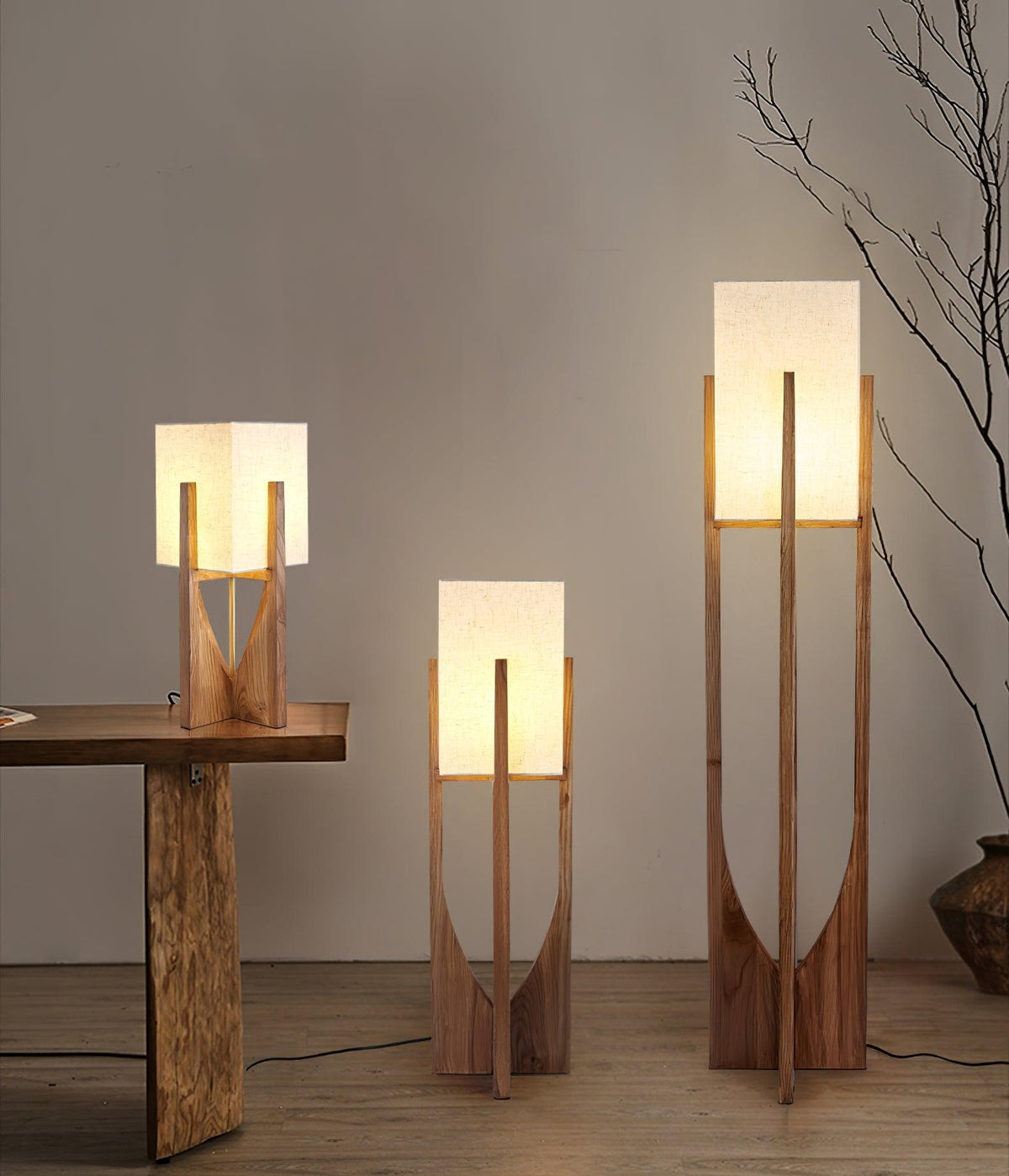 Three sizes of this Clérachic wood lamp stand together on a bare floor, the tallest beside a bare branch and a rough wooden table.