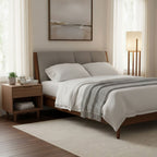 A Clérachic floor lamp stands behind a made bed with grey linen, beside a pale walnut bedside table and a framed landscape print.