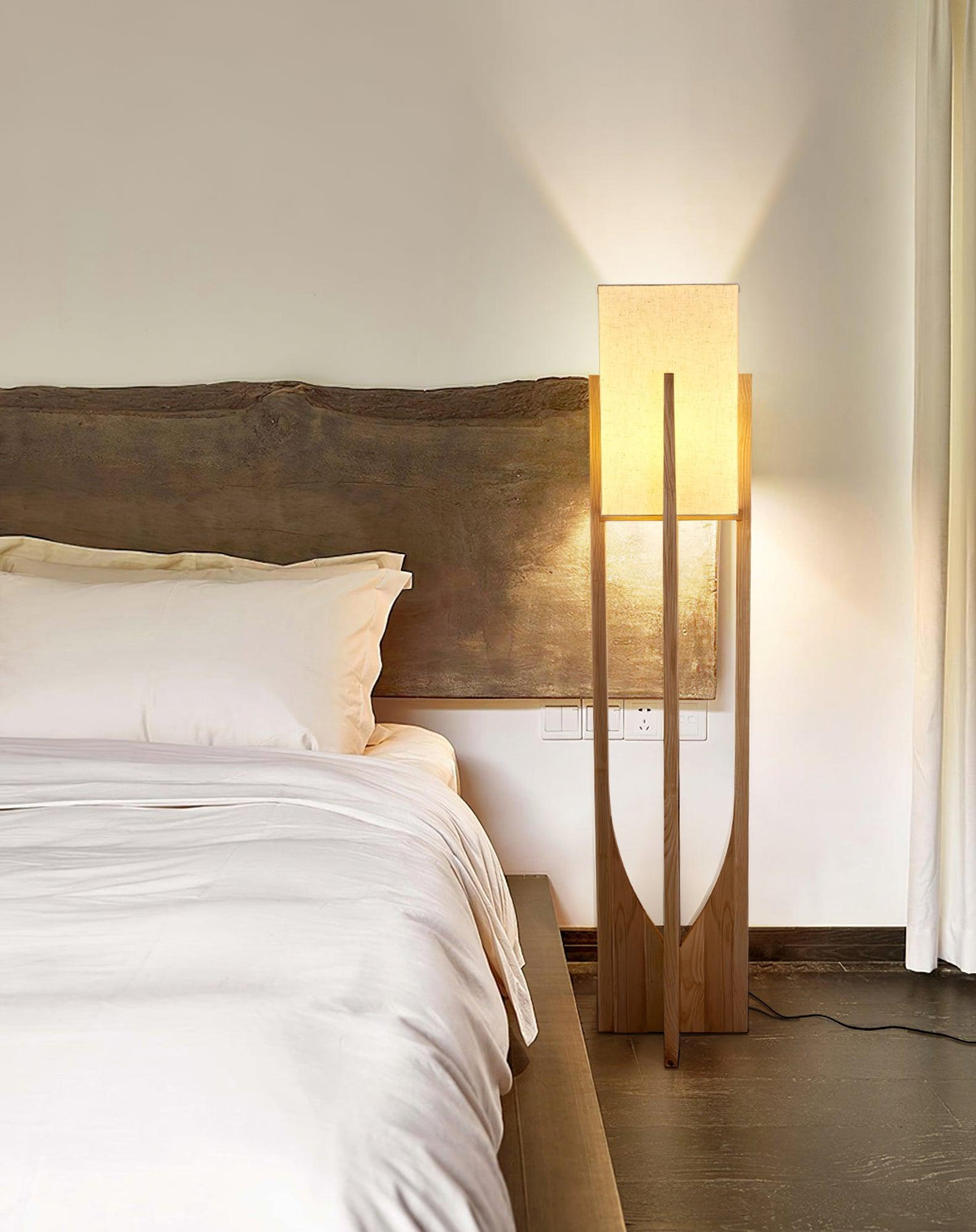 Floor lamp from Clérachic, its fabric shade lit, standing at a bedside by a live-edge wooden headboard and white bedding.