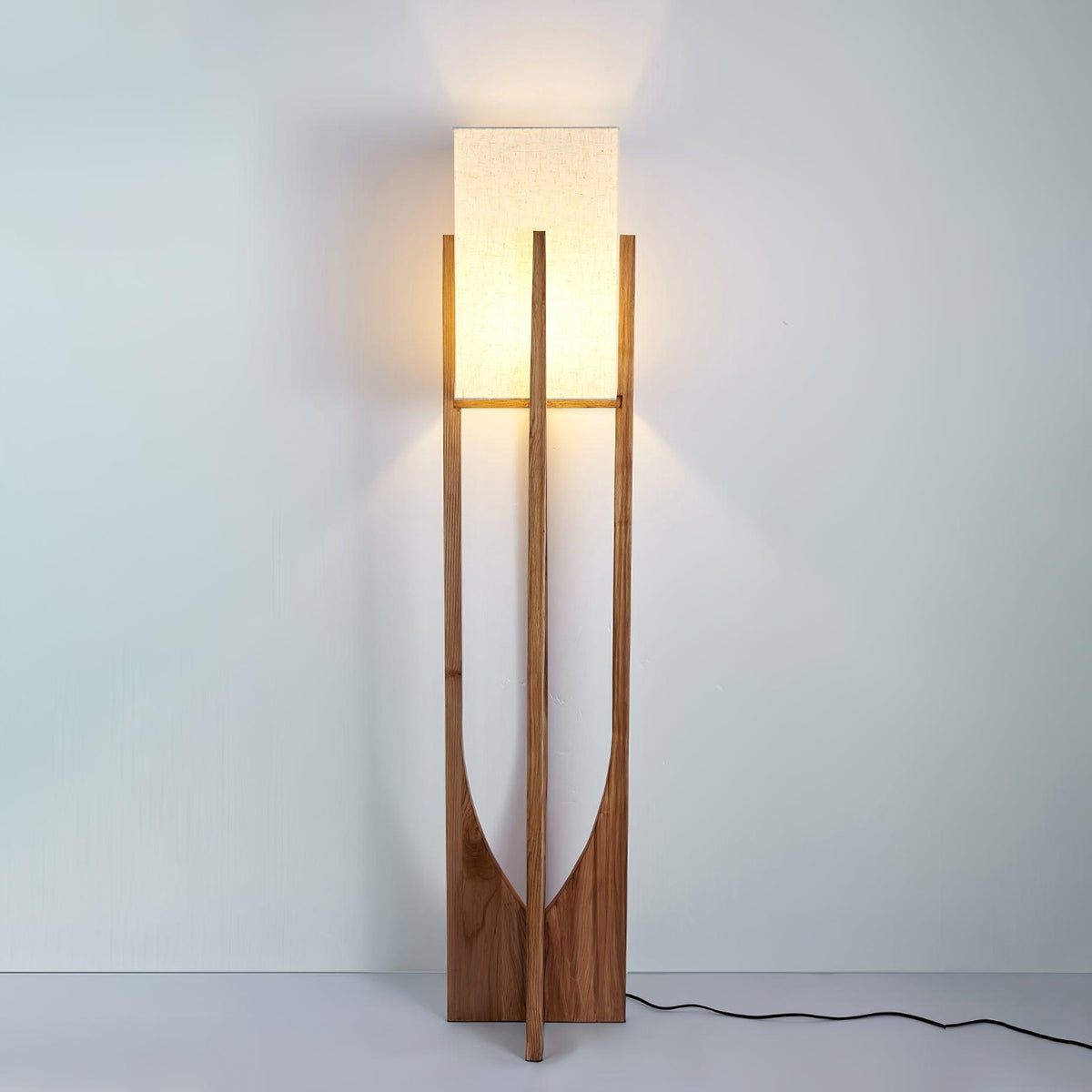 Lit Clérachic floor lamp against a pale blue wall, casting warm light on the wall above its fabric shade, with the black cord on the floor.
