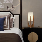 Small Clérachic wood lamp with a white fabric shade on a black lacquered cabinet beside a bed with cushions in navy and brown.