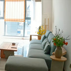 Small Clérachic floor lamp in the far corner of a living room with a blue-green sofa, a low table and a bright window.