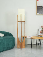 Clérachic natural wood floor lamp, unlit, with a white fabric shade, beside a green velvet sofa and a framed print.