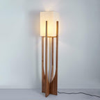 Lit Clérachic floor lamp in a dark wood frame, with a warm glow on the wall above and a black cord trailing on the floor.