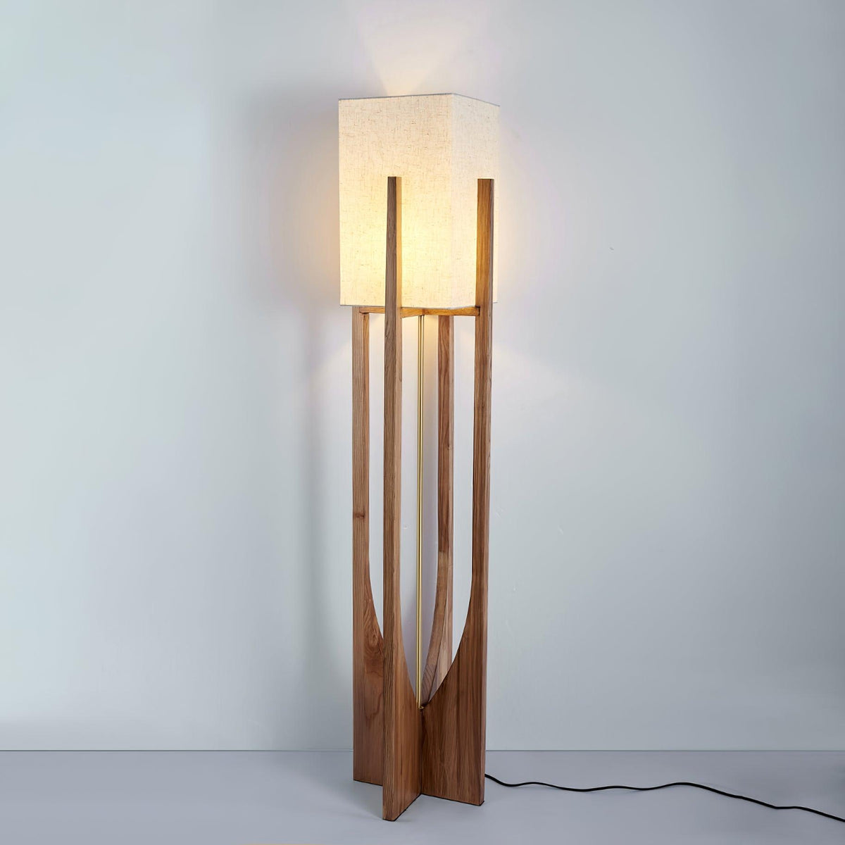 Lit Clérachic floor lamp in a dark wood frame, with a warm glow on the wall above and a black cord trailing on the floor.